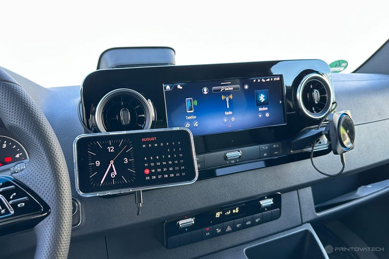 K&ouml;nnte beinhalten: Ein Armaturenbrett im Auto mit einem digitalen Display, das Radio- und Telefonoptionen anzeigt. Ein Smartphone zeigt eine Uhr und einen Kalender f&uuml;r August an. Ein kleiner Lautsprecher ist am Armaturenbrett montiert. Das Armaturenbrett ist schwarz mit silbernen Akzenten.