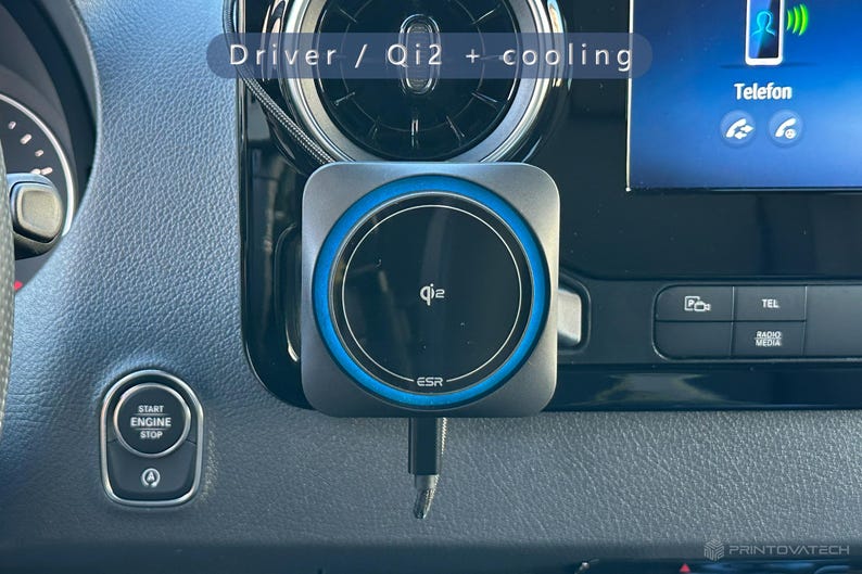 K&ouml;nnte beinhalten: Ein schwarzes und graues Qi2-Wireless-Ladeger&auml;t mit blauem Akzentring ist im Armaturenbrett eines Autos montiert. Das Ladeger&auml;t hat das ESR-Logo und den Text "Driver / Qi2 + cooling". Das Armaturenbrett des Autos verf&uuml;gt au&szlig;erdem &uuml;ber eine Start/Stopp-Taste und einen Bildschirm mit der Aufschrift "Telefon".
