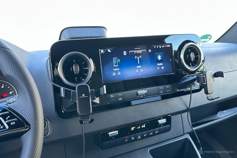K&ouml;nnte beinhalten: Armaturenbrett eines Fahrzeugs mit zentralem Bildschirm, der eine Benutzeroberfl&auml;che mit Symbolen f&uuml;r Telefon, Radio und Bluetooth anzeigt. Das schwarze Armaturenbrett umfasst L&uuml;ftungsschlitze und eine digitale Temperaturanzeige. Eine Telefonhalterung ist angebracht.