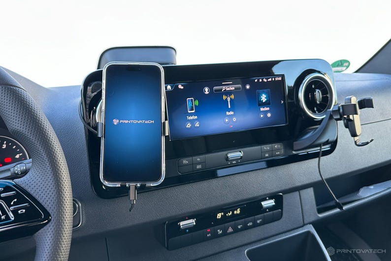 K&ouml;nnte beinhalten: Ein Armaturenbrett im Auto mit einem Smartphone in einer transparenten H&uuml;lle, das &uuml;ber ein Ladekabel angeschlossen ist. Das Telefon zeigt einen blauen Bildschirm mit dem Printovatech-Logo. Ein digitales Display zeigt Radio- und Bluetooth-Optionen. Das Armaturenbrett ist schwarz.