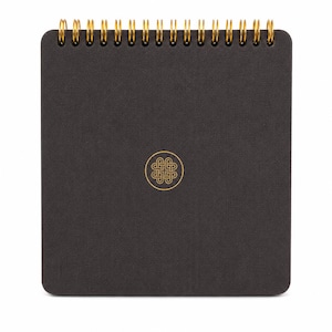 Puede incluir: Un cuaderno cuadrado con una cubierta texturizada de color gris carb&oacute;n oscuro y una encuadernaci&oacute;n de espiral met&aacute;lica dorada en la parte superior. El centro de la portada presenta un emblema circular dorado con un dise&ntilde;o geom&eacute;trico de nudo sim&eacute;trico.