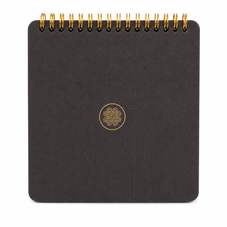 Puede incluir: Un cuaderno cuadrado con una cubierta texturizada de color gris carb&oacute;n oscuro y una encuadernaci&oacute;n de espiral met&aacute;lica dorada en la parte superior. El centro de la portada presenta un emblema circular dorado con un dise&ntilde;o geom&eacute;trico de nudo sim&eacute;trico.
