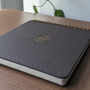 Puede incluir: Un cuaderno cuadrado con una cubierta de tela texturizada gris oscuro y una encuadernaci&oacute;n en espiral dorada. El centro de la portada presenta un emblema circular dorado en relieve con un intrincado dise&ntilde;o de nudo geom&eacute;trico.