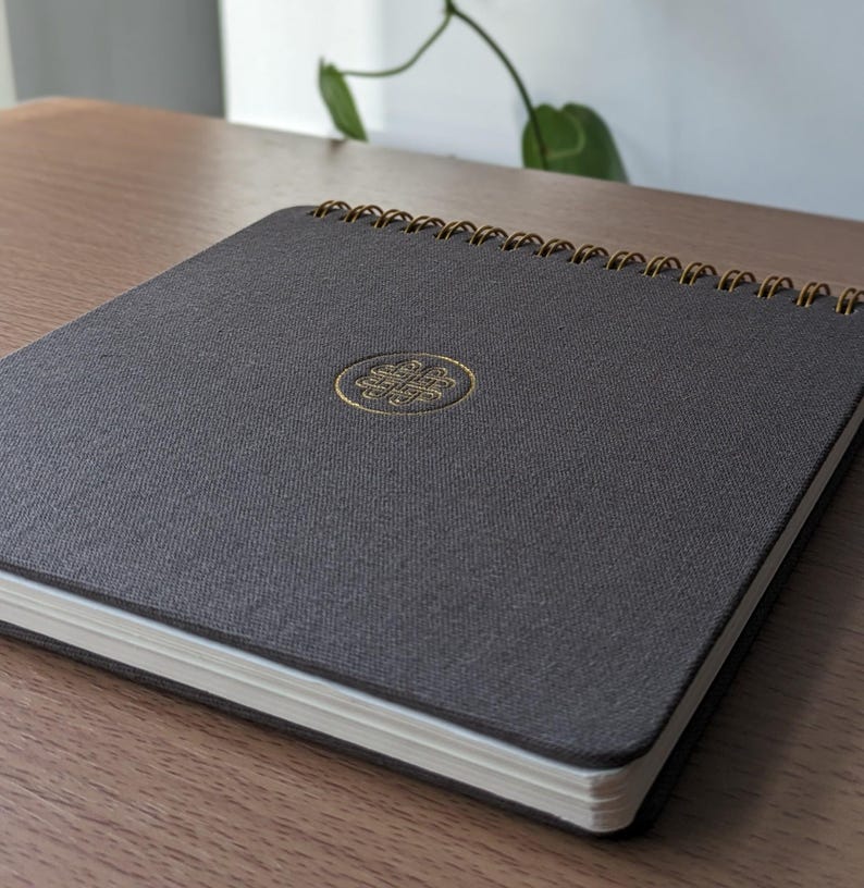 Puede incluir: Un cuaderno cuadrado con una cubierta de tela texturizada gris oscuro y una encuadernaci&oacute;n en espiral dorada. El centro de la portada presenta un emblema circular dorado en relieve con un intrincado dise&ntilde;o de nudo geom&eacute;trico.