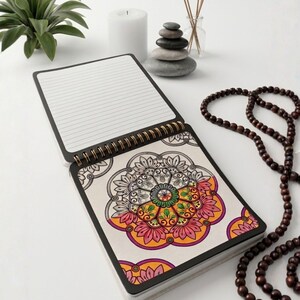 Puede incluir: Un cuaderno de espiral con una p&aacute;gina con l&iacute;neas horizontales y otra p&aacute;gina que muestra una colorida ilustraci&oacute;n de mandala con patrones florales en rosa, naranja, verde y amarillo, acentuada por un borde negro y encuadernaci&oacute;n dorada.