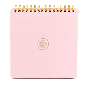 Puede incluir: Un cuaderno cuadrado con una cubierta texturizada de color rosa claro y una espiral dorada en la parte superior. El centro de la portada presenta un emblema circular dorado que contiene un dise&ntilde;o gr&aacute;fico complejo, sim&eacute;trico y entrelazado.