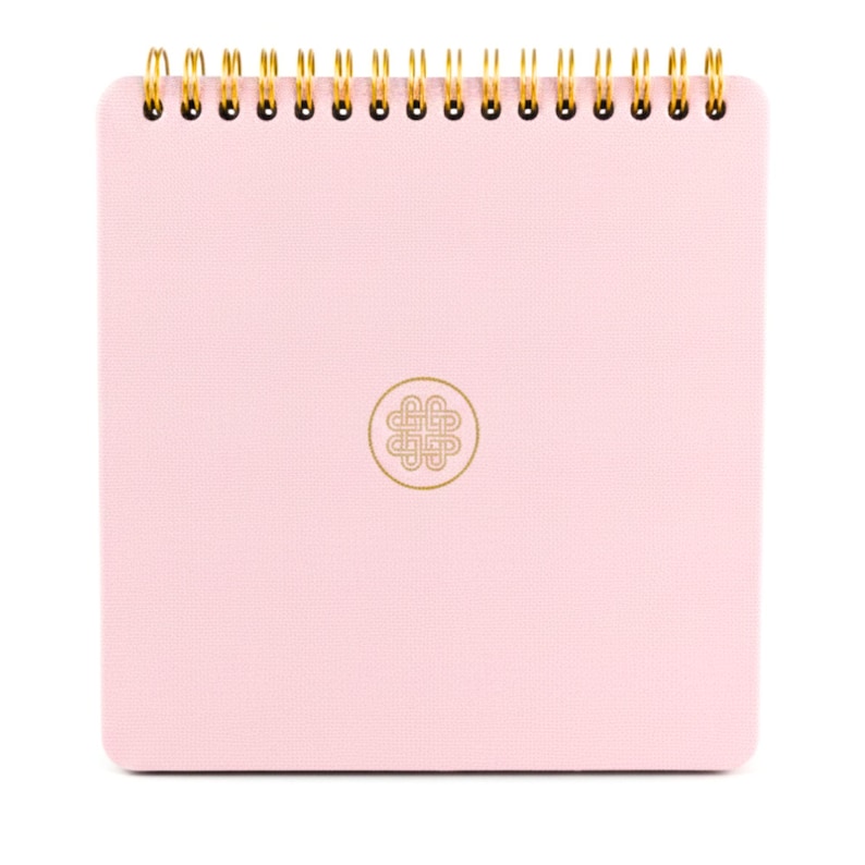 Puede incluir: Un cuaderno cuadrado con una cubierta texturizada de color rosa claro y una espiral dorada en la parte superior. El centro de la portada presenta un emblema circular dorado que contiene un dise&ntilde;o gr&aacute;fico complejo, sim&eacute;trico y entrelazado.