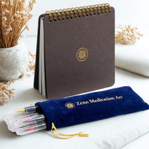Puede incluir: Un cuaderno de espiral marr&oacute;n oscuro con un emblema circular dorado en la portada, junto a una bolsa de terciopelo azul marino con cord&oacute;n que contiene varios pinceles de cuerpo transparente. La bolsa incluye el texto dorado Zenn Meditation Art.