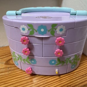 May include: A lavender jewelry box with a light blue handle and flower-shaped drawer pulls. The box is decorated with floral patterns in white, blue, and green. The box is made of plastic and has multiple drawers.