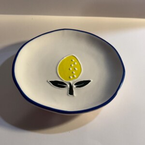 May include: A small, round, white ceramic dish with a blue rim. The dish features a yellow lemon design with black leaves and stem in the center. The lemon has white dots. The dish is likely used for serving small portions or as a decorative item.