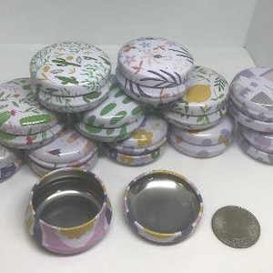 Op de afbeelding: Een verzameling kleine, ronde, metalen blikken met verschillende bloem- en geometrische patronen. De blikken hebben witte deksels en zijn op elkaar gestapeld.