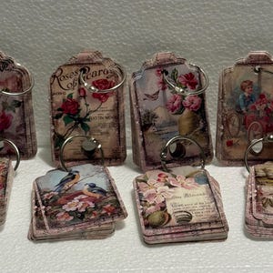 May include: A collection of vintage-style decorative tags with floral and pastoral imagery. Each tag features a metal ring and is adorned with roses, birds, and antique illustrations. The tags have a distressed, aged appearance.