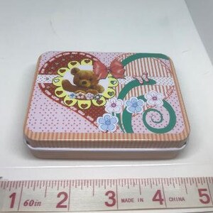 Puede incluir: Una caja de lata en forma de coraz&oacute;n rosa y marr&oacute;n con un borde blanco. La caja presenta un oso de dibujos animados con un lazo y flores. La caja mide aproximadamente 10 cm de ancho.