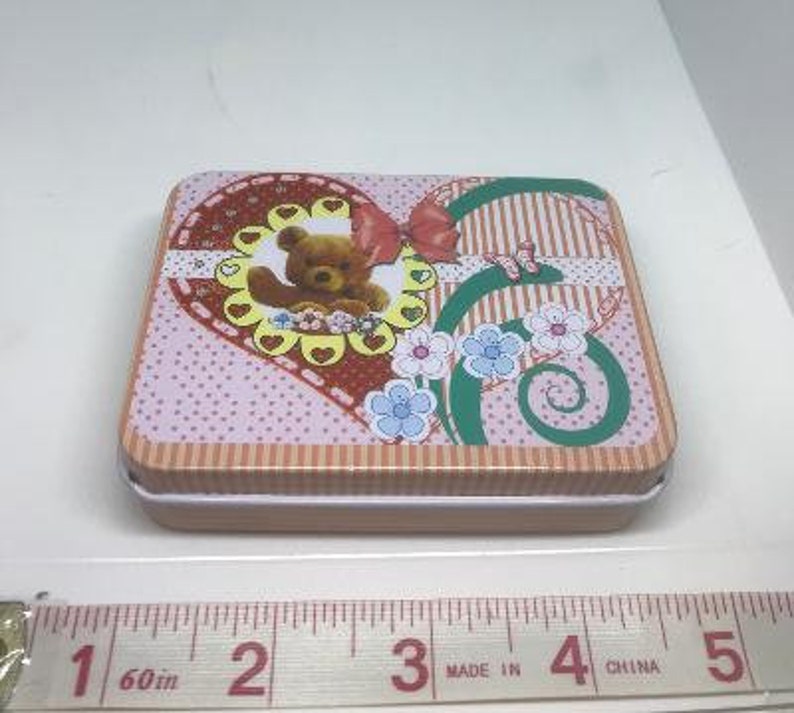 Puede incluir: Una caja de lata en forma de coraz&oacute;n rosa y marr&oacute;n con un borde blanco. La caja presenta un oso de dibujos animados con un lazo y flores. La caja mide aproximadamente 10 cm de ancho.