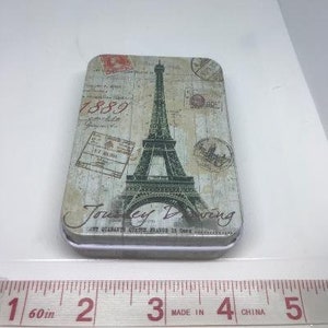 Puede incluir: Una caja de hojalata de estilo vintage con fondo blanco y una imagen en blanco y negro de la Torre Eiffel. El texto "Journey Dreaming" est&aacute; impreso en la caja, junto con el a&ntilde;o "1889".