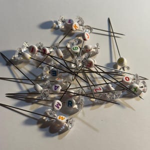 May include: A collection of sewing pins with clear, faceted heads. Each pin features a small, white bead with a single letter in various colors. The pins are scattered across a white surface, creating a craft supply arrangement.