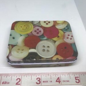 Puede incluir: Una peque&ntilde;a caja de metal rectangular con un patr&oacute;n colorido de botones. Los botones son de varios colores, incluyendo rojo, amarillo, azul, verde y blanco. La caja de metal es perfecta para guardar objetos peque&ntilde;os como joyas, botones u otras chucher&iacute;as.