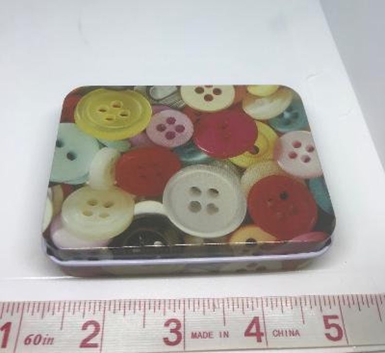 Puede incluir: Una peque&ntilde;a caja de metal rectangular con un patr&oacute;n colorido de botones. Los botones son de varios colores, incluyendo rojo, amarillo, azul, verde y blanco. La caja de metal es perfecta para guardar objetos peque&ntilde;os como joyas, botones u otras chucher&iacute;as.