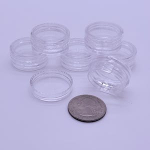 May include: A collection of small, clear, circular plastic containers with threaded rims, arranged in a cluster. A single United States quarter coin is positioned in the foreground to provide a visual scale for the size of the containers.