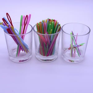 May include: Three clear glass tumblers containing assorted plastic yarn needles in vibrant colors including red, blue, green, yellow, and pink. The needles feature large eyes and are arranged in varying quantities across the three glass containers.