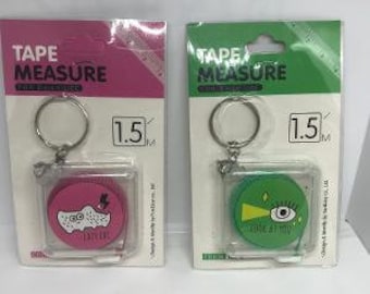 May include: Two 1.5 meter tape measures with keychains. One is pink with a white cartoon design and the other is green with a white eye design. Both have the text "TAPE MEASURE" on the packaging.