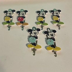 May include: Six wooden keychains featuring Mickey Mouse in different poses. Each keychain is painted in bright colors, including red, yellow, green, and blue. The keychains are attached to metal rings.