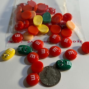 May include: A collection of colorful M&M's candies and charms, including red, yellow, green, and orange pieces. Some candies have the letter "m" printed on them. A US quarter coin is also present.