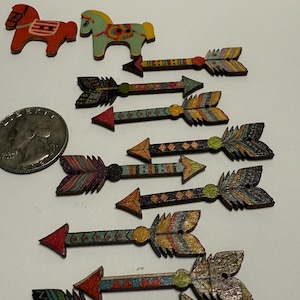 Puede incluir: Suministros de artesanía de madera con dos formas de caballo y varias formas de flecha. Las flechas tienen diseños de plumas coloridas y patrones geométricos. Los caballos son rojos y verdes con detalles pintados. Se incluye una moneda como referencia de tamaño.
