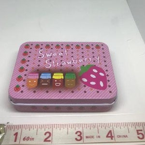 Puede incluir: Lata rectangular rosa con tapa blanca. La lata tiene un patr&oacute;n de rayas rosa y blancas y presenta una fresa con semillas blancas y cuatro tarros con caras sonrientes. El texto "Sweet Strawberry" est&aacute; impreso en la lata.