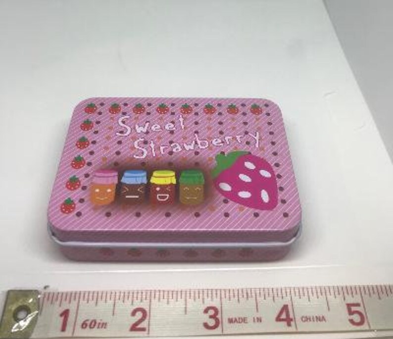 Puede incluir: Lata rectangular rosa con tapa blanca. La lata tiene un patr&oacute;n de rayas rosa y blancas y presenta una fresa con semillas blancas y cuatro tarros con caras sonrientes. El texto "Sweet Strawberry" est&aacute; impreso en la lata.