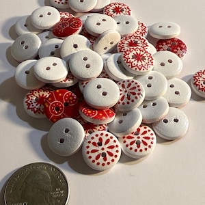 Puede incluir: Una colección de botones decorativos blancos y rojos. Los botones redondos con dos agujeros presentan diseños florales y de copos de nieve rojos. Se incluye una moneda de veinticinco centavos estadounidense como referencia de tamaño.