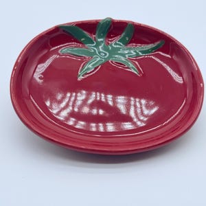 May include: A glossy, oval-shaped ceramic dish in a vibrant red color, featuring a raised, dark green stem and leaf design in the center. The surface is smooth and reflective, mimicking the appearance of a ripe tomato with a raised rim.