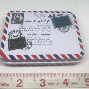Puede incluir: Una caja de metal blanca con rayas rojas y azules. La caja tiene un dise&ntilde;o de avi&oacute;n vintage y sello de correos con el texto "saves the day" y "very few are genuine".