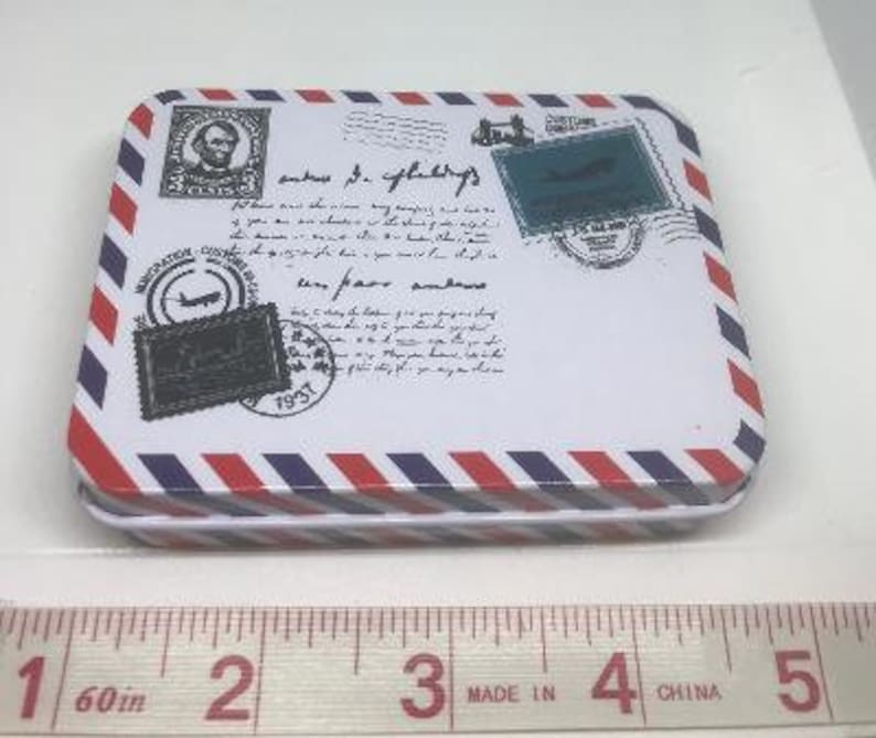 Puede incluir: Una caja de metal blanca con rayas rojas y azules. La caja tiene un dise&ntilde;o de avi&oacute;n vintage y sello de correos con el texto "saves the day" y "very few are genuine".