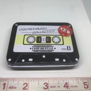 Puede incluir: Una lata negra en forma de casete con una etiqueta amarilla. La etiqueta dice "Country Music Collection" y "New". La lata tiene un dise&ntilde;o en blanco y negro con las palabras "Confidential" y "Side B".