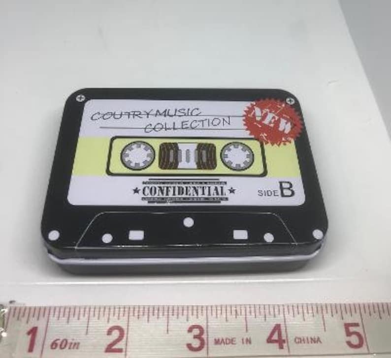 Puede incluir: Una lata negra en forma de casete con una etiqueta amarilla. La etiqueta dice "Country Music Collection" y "New". La lata tiene un dise&ntilde;o en blanco y negro con las palabras "Confidential" y "Side B".
