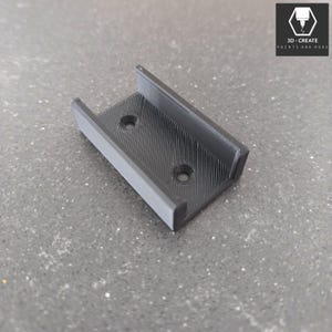 Könnte beinhalten: Eine schwarze, 3D-gedruckte Halterung mit strukturierter Oberfläche und zwei kreisförmigen Löchern. Die Halterung hat eine rechteckige Basis mit zwei erhöhten Seiten. Das Logo "3D-CREATE PRINTS AND MORE" befindet sich in der oberen rechten Ecke.