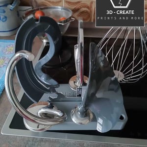 Könnte beinhalten: Küchengeräte-Aufsätze, darunter ein Knethaken, ein Rührbesen und ein Schneebesen, werden auf einer Arbeitsplatte präsentiert. Die Aufsätze sind grau und silberfarben, mit einem Schneebesen aus Edelstahl im Hintergrund. Der Text "3D - CREATE PRINTS AND MORE" ist sichtbar.