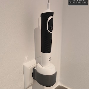 Könnte beinhalten: Ein 3D-gedruckter Wandhalter für eine elektrische Zahnbürste, gedruckt aus grauem Kunststoff. Der Halter ist an einer normalen Steckdose befestigt und hält die Zahnbürste in vertikaler Position.