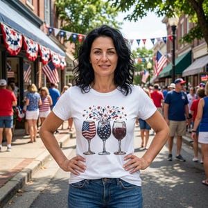 May include: A white short-sleeved t-shirt featuring a graphic print of three wine glasses with patriotic designs, including an American flag pattern, a blue field with white stars, and a solid red wine fill, topped with red and blue star bursts.