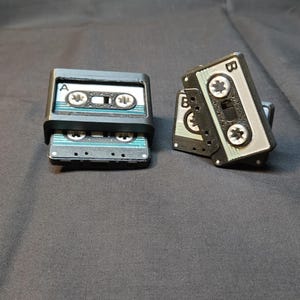 May include: Three miniature cassette tape replicas in black and gray. One set of two tapes is stacked, and another is leaning against the stack. The tapes have labels with the letters "A" and "B" and a retro design.