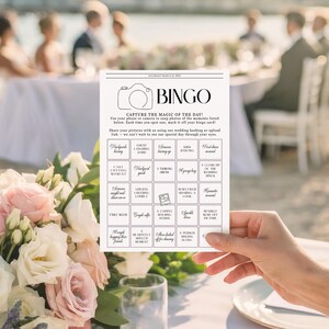 May include: A white rectangular wedding bingo card with black text and a camera icon. The card features a grid of squares with prompts like 'Newlyweds kissing' and 'Cake cutting moment'. The text 'SATURDAY MARCH 21, 2025' is printed at the top.