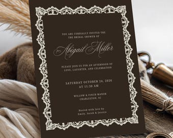 May include: A dark brown rectangular invitation card featuring a cream-colored intricate lace border. The text reads: You are cordially invited the bridal shower of Abigail Miller, please join us for an afternoon of love, laughter, and celebration, Saturday October 24, 2026 at 11:30 AM, Willow & Finch Manor Charleston, SC, hosted with love by Emily, Sarah & Jessica.