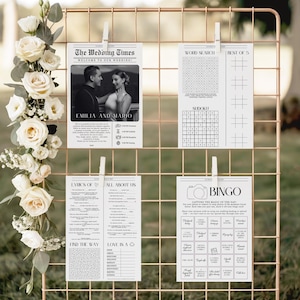 May include: A rose gold wire grid display holds four white wedding activity cards clipped with white clothespins. The cards feature black text and graphics, including a photo of a couple, a word search, a sudoku puzzle, a bingo game, and lyrics.