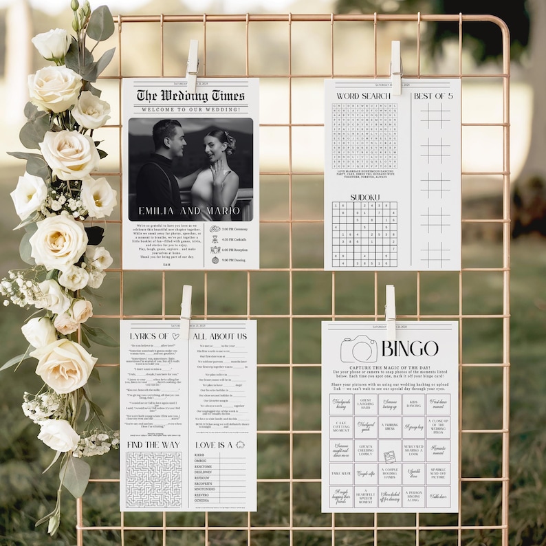 May include: A rose gold wire grid display holds four white wedding activity cards clipped with white clothespins. The cards feature black text and graphics, including a photo of a couple, a word search, a sudoku puzzle, a bingo game, and lyrics.