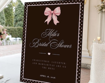 May include: A rectangular sign with a dark brown background and a white polka dot border. It features a light pink bow at the top, the text "Mila's Bridal Shower", a small heart, the text "Charcuterie, laughter, and margaritas are on the menu", and "April 18, 2026".