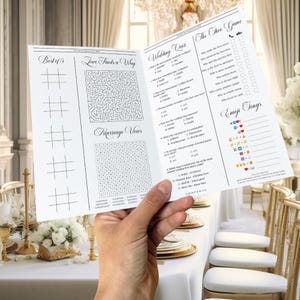 May include: A white wedding game booklet with various games and quizzes, including tic-tac-toe, a maze, and a word search. The booklet also features a wedding quiz and emoji song game. The booklet is held in a person's hand.
