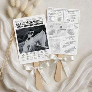 May include: Two wedding fans with wooden handles and white ribbon bows. The fans are printed with a newspaper-style design. One fan features a black and white photo of a couple, the text "The Wedding Gazette," and a wedding timeline. The other fan includes wedding party details and a word search.