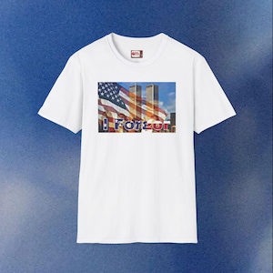 Op de afbeelding: Wit T-shirt met een afbeelding van de Amerikaanse vlag en de Twin Towers, met het woord "Forever" eronder gedrukt. Het shirt heeft een ronde hals en korte mouwen.
