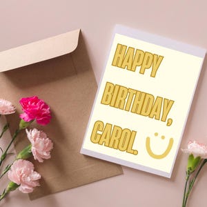 May include: A birthday card with the text "HAPPY BIRTHDAY, CAROL" in gold lettering, accompanied by a smiling face. The card is next to a brown envelope and pink flowers.